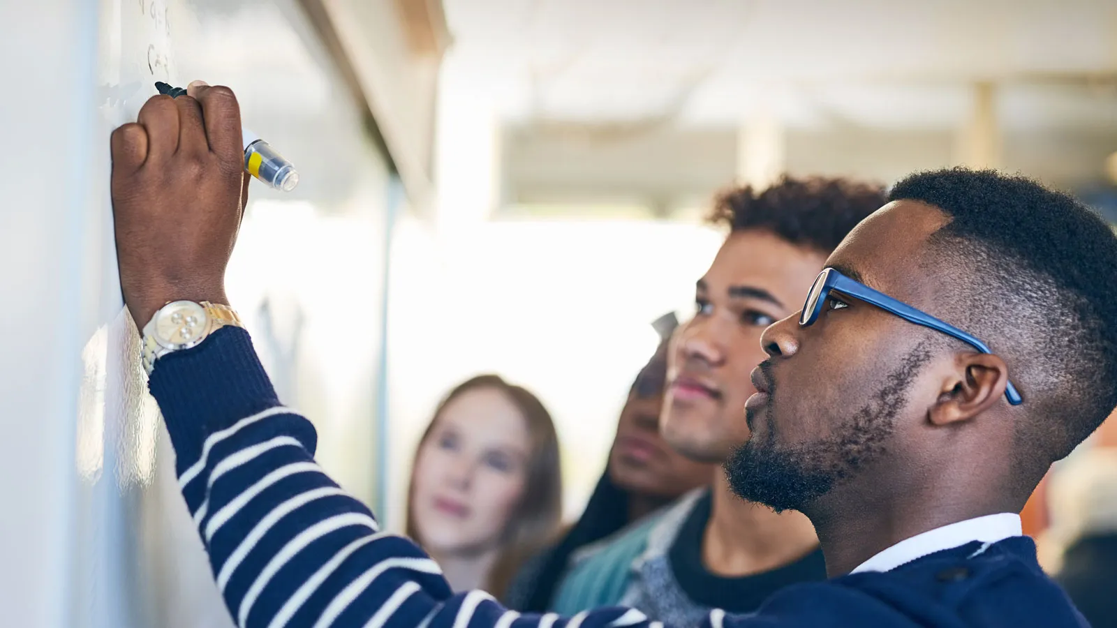 African American male college student writing on a whiteboard and collaborating with fellow students